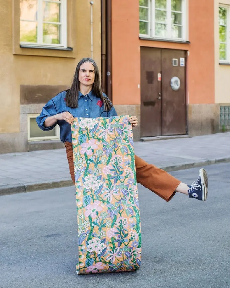 Цифровые обои для стен Hanna Werning Friviva Orange, арт. 9841W от Borastapeter (Швеция) – экзотические растения переплетаются с полевыми цветами из шведской флоры, создавая буйный, многослойный и невероятно детализированный узор в ярких оттенках розового, фиолетового, синего, зелёного и белого на светящемся приглушённо-оранжевом фоне. Купить обои в интернет-магазине Odesign с бесплатной доставкой, чтобы создать стильный интерьер для комфортной жизни в скандинавском стиле. Фото обоев с автором.