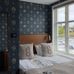 Уютная спальня с дизайнерскими обоями Ängsflora Midnight Teal (арт. 6952) из коллекции Österlen. На глубоком тёмно-синем фоне — многоцветные букеты луговых цветов: голубые колокольчики, лесные анемоны, льнянки, незабудки, примулы и земляника, нарисованные вручную дизайнером Нуми Спанж. В интерьере — кровать с деревянным изголовьем и белым постельным бельём, льняные подушки с вышитыми буквами G, латунное настенное бра, белая римская штора, окно с видом на воду и лодку, светлые деревянные полы. Атмосфера — спокойная, атмосфера залитого лунным светом шведского сада.