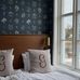 Уютная спальня с дизайнерскими обоями Ängsflora Midnight Teal (арт. 6952) из коллекции Österlen. На глубоком тёмно-синем фоне — многоцветные букеты луговых цветов: голубые колокольчики, лесные анемоны, льнянки, незабудки, примулы и земляника, нарисованные вручную дизайнером Нуми Спанж. В интерьере — кровать с деревянным изголовьем и белым постельным бельём, льняные подушки с вышитыми буквами G, латунное настенное бра, белая римская штора, окно с видом на воду и лодку, светлые деревянные полы. Атмосфера — спокойная, атмосфера залитого лунным светом шведского сада.