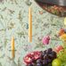 Обои с птицами и цветами Hummingbirds Duck Egg Mica, арт 124/1007 в винтажном стиле на мятно-зеленом фоне. Праздничный натюрморт: желтые свечи в стеклянных подсвечниках, букет роз, виноград, гранат и лайм на столе. Ботанический узор для гостиной и столовой.