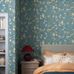 Интерьер спальни с флизелиновыми обоями Boråstapeter Sophia Magdalena Sky Blue (арт. 8706). Узор шинуазри с цветущими ветвями, экзотическими птицами и бабочками в терракотовых и бледно-розовых тонах в обрамлении светло-зелёной листвы на приглушённом бирюзово-голубом фоне. Уютная усадебная обстановка с кроватью, тумбочкой, красной лампой и подушками. Коллекция Herrgård от Boråstapeter.