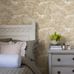 Интерьер спальни с флизелиновыми обоями Boråstapeter Alnarp Beige (арт. 8716). Пышный пейзажный узор с деревьями, рекой и мостом в тёплой бежевой палитре. Уютная усадебная обстановка с кроватью с резным изголовьем, тумбочкой, цветами и подушками. Коллекция Herrgård от Boråstapeter.