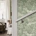 Интерьер коридора с флизелиновыми обоями Boråstapeter Alnarp Green (арт. 8717). Пышный пейзажный узор с деревьями, рекой и мостом в мягкой приглушённой светло-зелёной палитре. Уютная усадебная обстановка с белыми перилами, диваном, пуфом и видом в гостиную. Коллекция Herrgård от Boråstapeter.