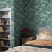 Интерьер спальни с флизелиновыми обоями Boråstapeter Alnarp Blue&Green (арт. 8718). Пышный пейзажный узор с деревьями, рекой и мостом в насыщенной сине-зелёной палитре. Уютная усадебная обстановка с кроватью, стеллажом, красной лампой и подушками. Коллекция Herrgård от Boråstapeter.